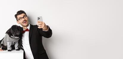 Cheerful dog owner in suit celebrating Christmas with dog, taking selfie on smartphone and hugging cute black pug in costume, white background photo