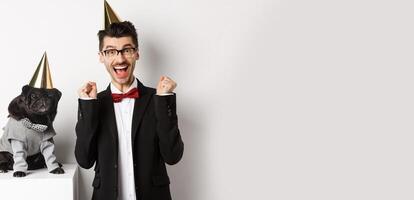 Cheerful young man shouting for joy, dog and owner wearing birthday party cones and celebrating, guy rejoicing and staring at camera, white background photo