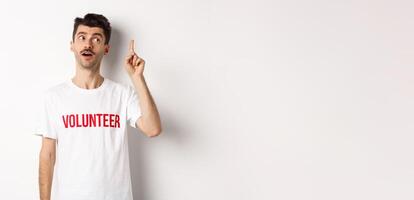Handsome young man in volunteer t-shirt having an idea, raising finger and saying suggestion, white background photo