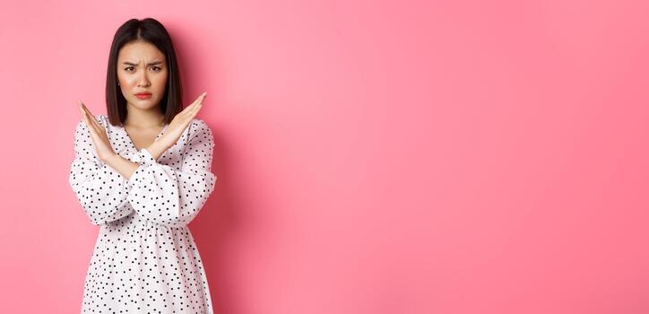 enojado asiático mujer contar detener, haciendo cruzar a prohibir o desaprobar algo, ceñudo disgustado, en pie terminado rosado antecedentes foto
