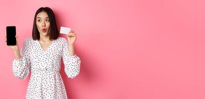 Cute asian woman shopping online, showing bank credit card and mobile screen, smiling and looking left at copy space, standing over pink background photo