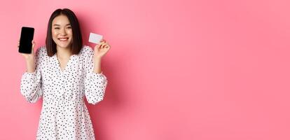 Joyful korean woman showing smartphone screen and credit card, paying for internet order, demonstrating online shopping app, standing over pink background photo