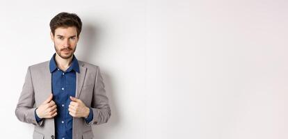 Confident young man with beard fixing suit and looking sassy at camera, feeling ready and determined, standing on white background photo