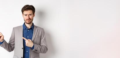 Bad idea. Displeased skeptical guy in business suit smirk and frown, pointing fingers left at upsetting news, looking hesitant at camera, white background photo