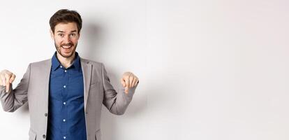 Hey check this out. Happy businessman in suit pointing fingers down and smiling, inviting to event, showing link, standing on white background photo
