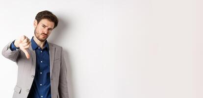 Negative emotions. Disappointed businessman frowning, showing thumb down to disapprove product, dislike something bad, stading on white background photo