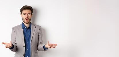 Confused and shocked businessman raising hands up and asking what happened, frowning disappointed, waiting for explanation, standing in suit on white background photo