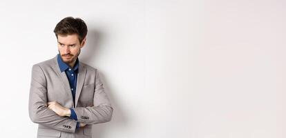 Moody guy in suit looking aside with unfair face, cross arms on chest and frowning angry, standing upset against white background photo