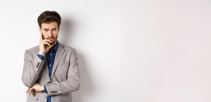 Troubled frowning business man in suit looking at camera pensive, thinking or making decision, standing on white background photo