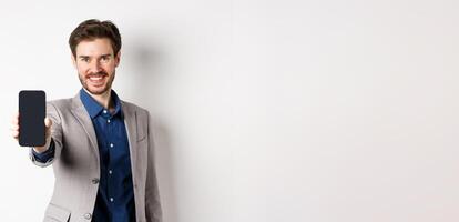 E-commerce and online shopping concept. Confident businessman in suit stretch out hand with empty smartphone screen, showing on phone, standing against white background photo