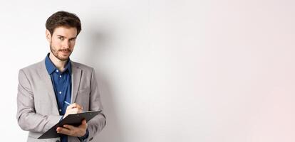 Successful businessman in grey suit taking notes, working in office clothing, holding clipboard and looking at camera, white background photo