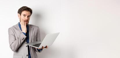 Shocked and troubled businessman have trouble on laptop, looking worried at camera, made mistake at work, standing on white background in suit photo
