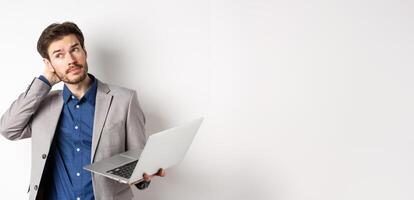 Thoughtful businessman scratch head and look away, making decision while working on laptop, standing against white background photo