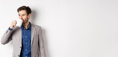 Handsome businessman in suit drinking coffee or tea from office mug, looking aside at logo, standing against white background photo