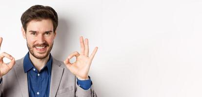 Smiling businessman in suit showing okay signs in approval, guarantee quality product, praise good thing, standing on white background photo