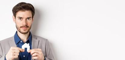 Real estate and insurance concept. Salesman realtor showing small paper house cutout and looking at camera, selling property, white background photo