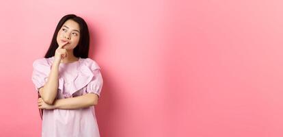 Pensive smiling asian girl making choice, looking at upper left corner logo and thinking, having an idea, standing cunning on pink background photo