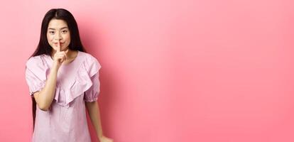 Cute asian girl hiding secret, hushing with finger pressed to lips and smiling, asking to keep quiet, standing in dress on pink background photo