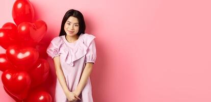 Cute asian girl in dress looking shy and smiling, standing modest near valentines day balloons, blushing on romantic date, looking at camera, pink background photo