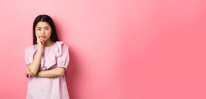Bored asian teen girl look indifferent at camera, lean face on hand in skeptical pose, standing reluctant against pink background photo