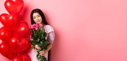 Valentines day concept. Tender and gentle teenage girl holding romantic flowers, standing near red heart balloons from lover and gazing with love, standing on pink background photo