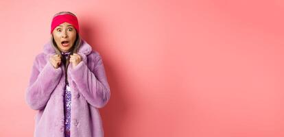 Fashion and shopping concept. Scared elderly woman scream and staring startled at camera, jumping from fear, wearing purple winter coat and headband, pink background photo