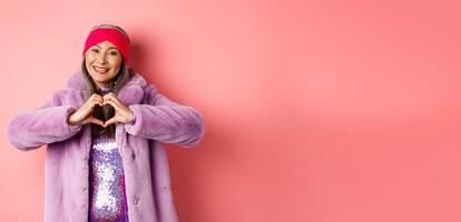 Romance and valentines day. Happy asian senior woman showing heart sign, I love you gesture, smiling and looking caring at camera, standing in faux fur coat, pink background photo