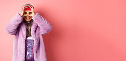 Fashion and shopping. Happy asian grandmother in stylish faur fur coat, looking through hand glasses, making binoculars and smilinga t camera, standing over pink background photo