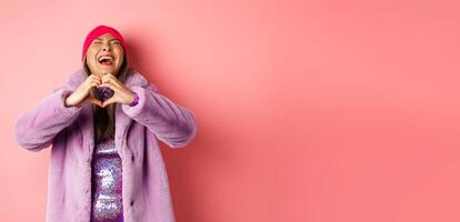 Valentines day and shopping concept. Happy asian woman in stylish outfit showing heart sign and laughing carefree, standing in faux fur against pink background photo