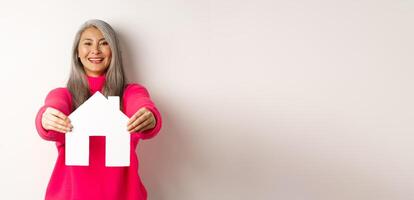 Real estate. Happy korean middle-aged woman with grey hair, showing paper house model and smiling, standing in pink sweater over white background photo