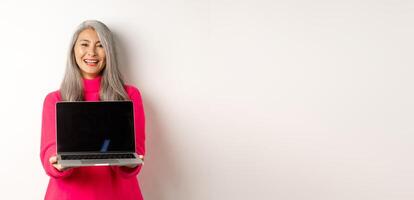 E-commerce concept. Beautiful asian senior businesswoman showing blank laptop screen and smiling, standing in pink sweater over white background photo