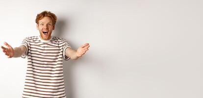 Cheerful man with red curly hair reaching hands forward, stretch out arms to welcome or congratulate you, smiling happy, standing over white background photo
