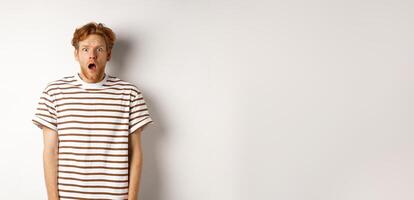 Shocked and scared redhead man screaming, open mouth and staring terrified at camera, standing over white background photo