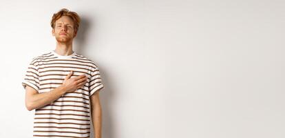 Young bearded redhead man holding hand on heart and looking patriotic, standing during national anthem, standing over white background photo