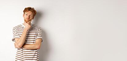 Portrait of thoughtful man with red hair thinking, looking at upper left corner and making choice, standing over white background photo