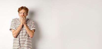 Shocked redhead guy gossiping, giggle in hands and looking left impressed, standing over white background photo