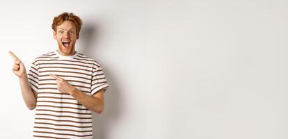 Cheerful man with red hair checking out promo offfer, pointing left at logo and smiling at camera, standing over white background photo