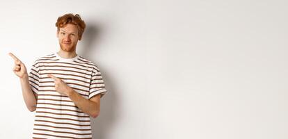 Doubtful redhead guy pointing fingers left at looking skeptical at camera, standing over white background photo