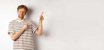 Judgemental redhead guy frowning disappointed, pointing fingers upper right corner at something bad, white background photo