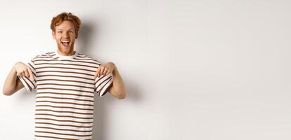 Enthusiastic young man with ginger hair and bristle showing advertisement, pointing fingers down and smiling, white background photo