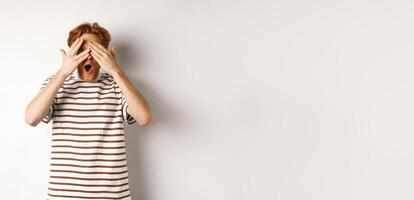Shocked and embarrassed redhead guy covering eyes, peeking through fingers and gasping, standing over white background photo