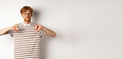 Disappointed young man with red hair and glasses disagree, showing thumbs-down and frowning displeased, standing over white background photo