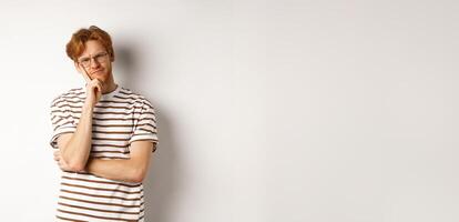 Young man with red messy hair and glasses looking doubtful, thinking and squinting at camera, standing thoughtful over white background photo