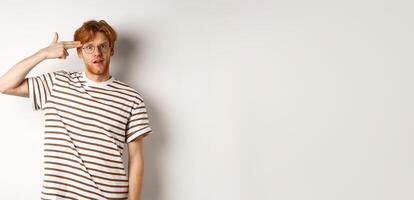 Annoyed teenage redhead guy making finger gun near head, staring bothered and irritated at camera, white background photo