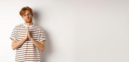 Silly redhead man asking for help, holding hands in pleading gesture and pucker lips, say please while begging you, standing over white background photo