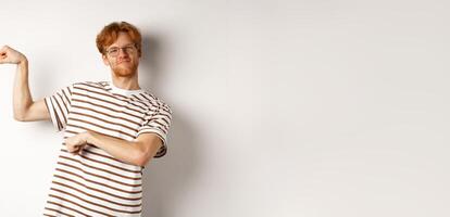 Image of confident and strong redhead man flexing biceps, showing muscles after gym, standing over white background photo