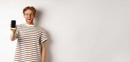Technology and e-commerce concept. Happy young redhead man in glasses showing blank smartphone screen, looking at camera amazed, standing over white background photo