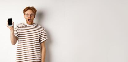 Technology and e-commerce concept. Surprised and shocked redhead guy checking out online promotion, showing blank smartphone screen and drop jaw, white background photo