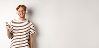 Technology concept. Young man with red hair and beard listening music in headphones and using smartphone, smiling at camera, white background photo