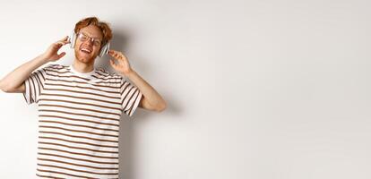 Technology concept. Happy redhead man listening music in headphones and singing along, standing over white background photo